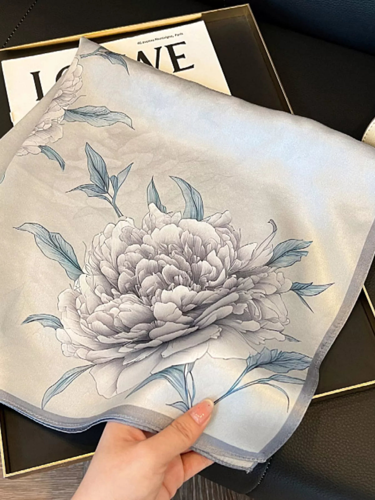 Hand holding the light gray scarf open to show a large floral motif and gray border.