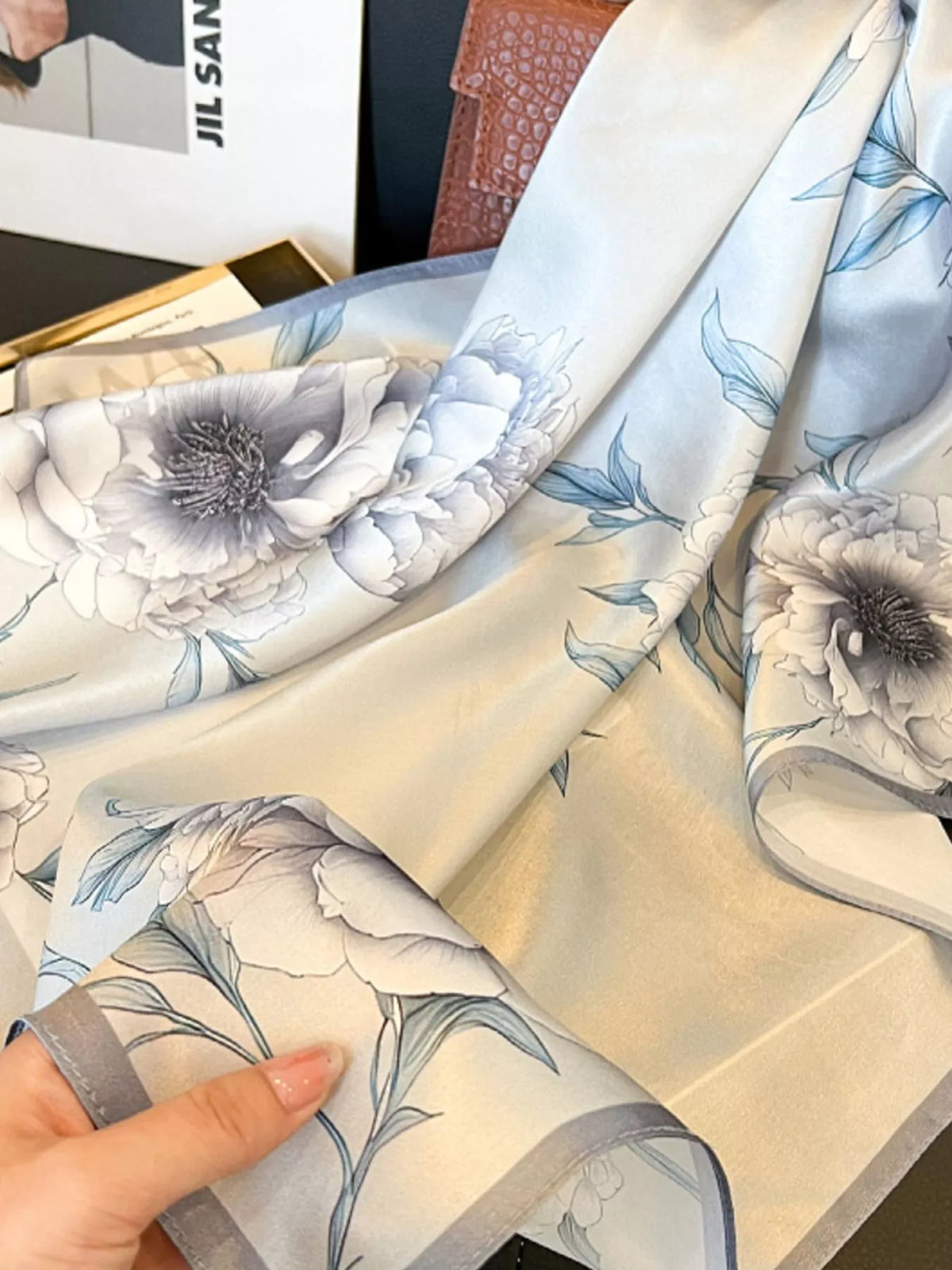 Light gray floral scarf draped beside a brown handbag with blue-toned leaf details.