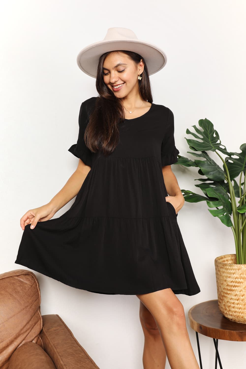 "Model lifting the hem of the black Mandy tiered V-neck mini dress to show the tiered skirt and pocket detail."